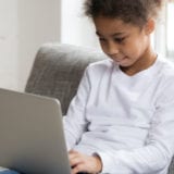 Curious preschool african mixed race girl using laptop on couch, little smart black kid typing on computer chatting with friends online alone at home, child security, children and gadget concept