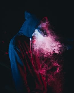 Hooded teenager in front of black background standing amidst pink vapor from e-cigarette