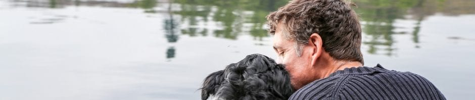 Behind view of man and dog sitting and hugging in front of lake