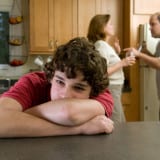 Divorce - adolescent leaning on table with head in arms, with fighting parents in the background